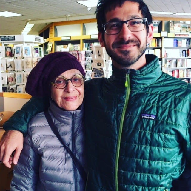 woman in grey jacket and man with green jacket with arm around her in bookstore