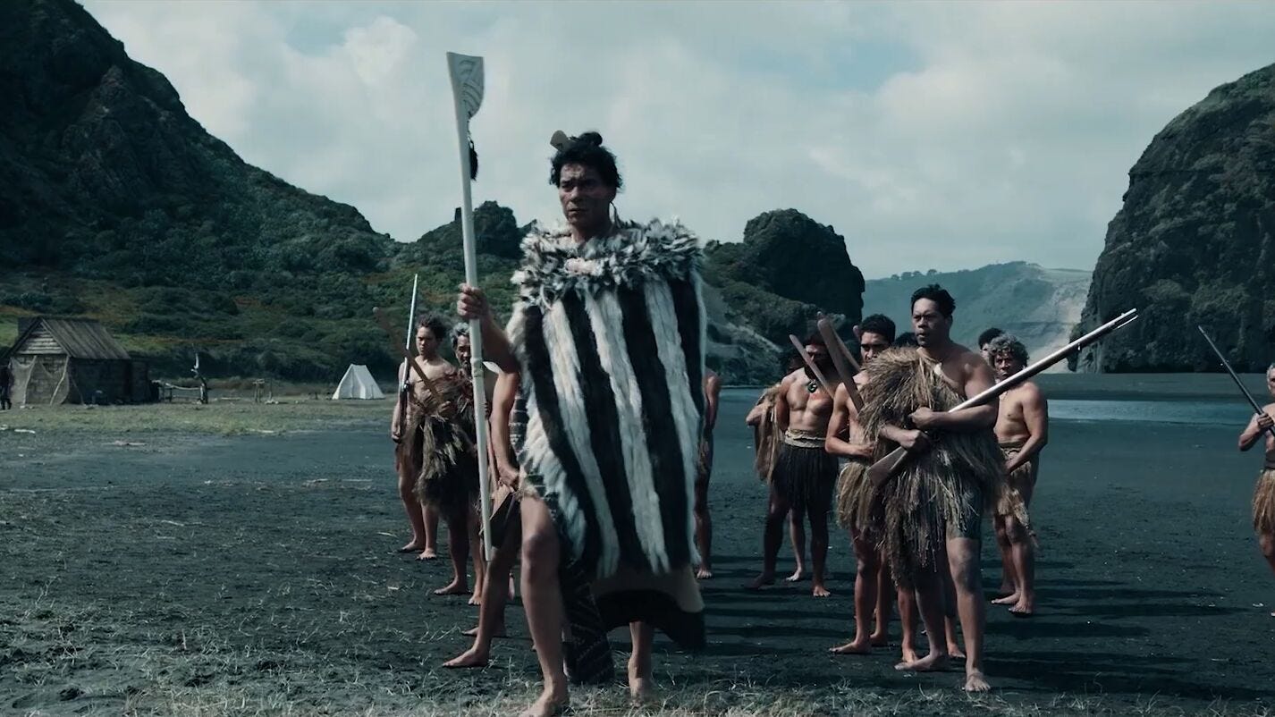 A group of Māori warriors dress in pre-colonial clothes, standing on a beach with taiaha and muskets. At their front is Maianui (Antonio Te Maioha)