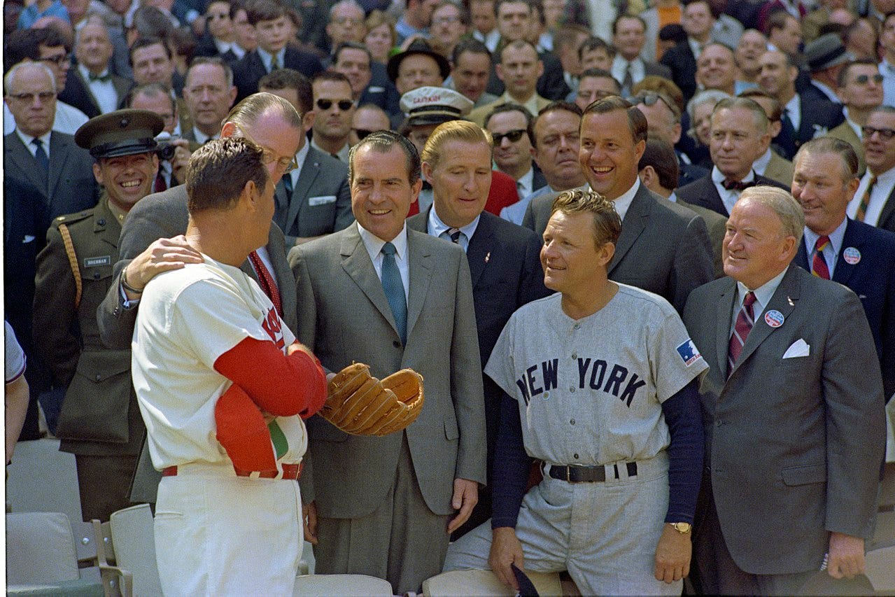 President Richard Nixon wears a baseball mitt while talking with the managers of the Washington Senators and the New York Yankees in front of a crowd of baseball fans.