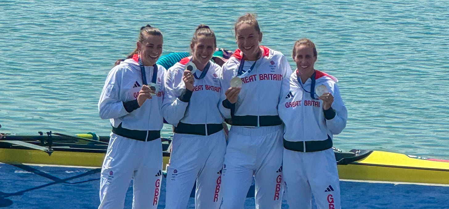 Four women in Team GB Olympic tracksuits stand on a podium in front of a lake holding their silver medals