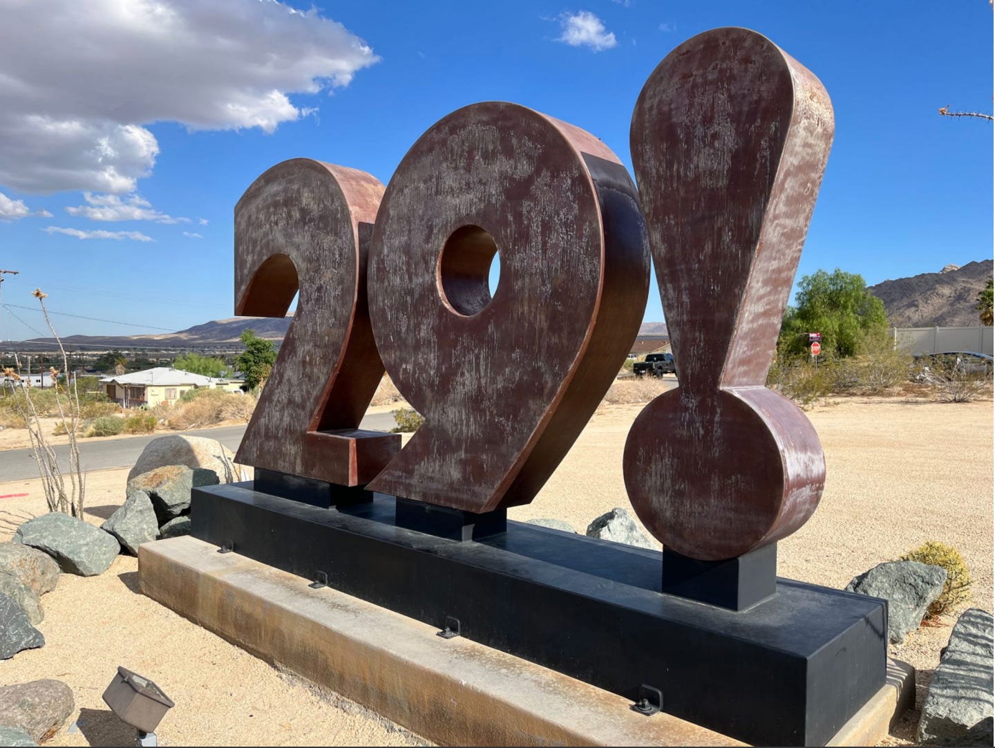 Close up of a brown sculpture consisting of the numbers 2 and 9 with an exclamation point, on a brown base. The sculpture is outside in a sandy lot in the desert and is by Chuck Caplinger.