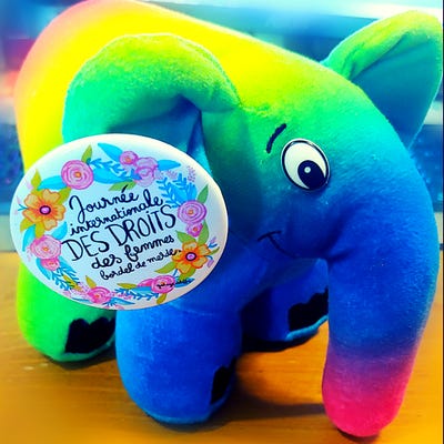 photo d'une peluche éléphant mascote de php (elePHPant) aux couleurs arc en ciel sur laquelle est épinglée un badge qui dit "Journée internationale DES DROITS des femmes bordel de merde" entouré d'une couronne fleuries.