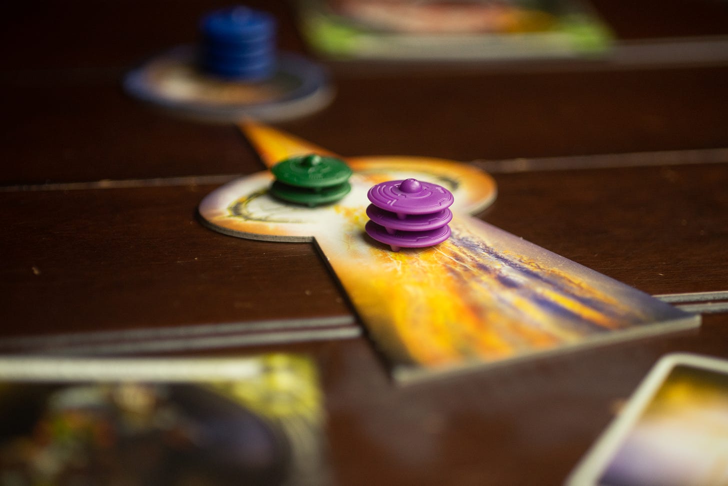Ships from the board game Cosmic Encounter placed on a piece indicating a player's planet being attacked. There are three purple pieces and two green pieces. In the background and out of focus, there are four blue ships defending the attacked planet.