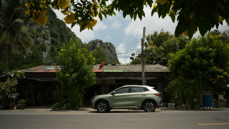 A photo of the BYD Atto 3 car parked in front of a small shop near a cliff taken across the street with yellow flowers and plants framing the top of the image.