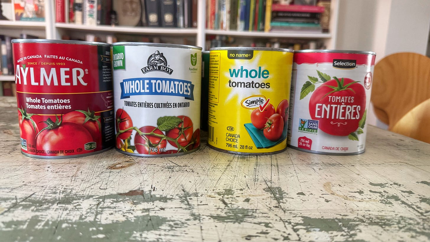 Cans of tomatoes on a dining room table. From left to right: Aylmer Whole Tomatoes, Farm Boy Whole Tomatoes, No Name Whole Tomatoes, Selection Tomates Entieres (whoops had the French side showing)