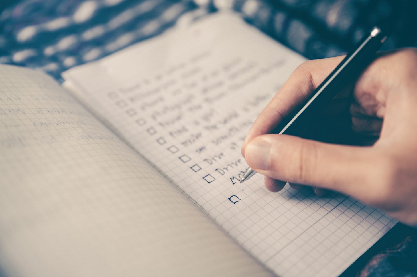 A hand holding a pen writing a to-do list in a small graph paper notebook