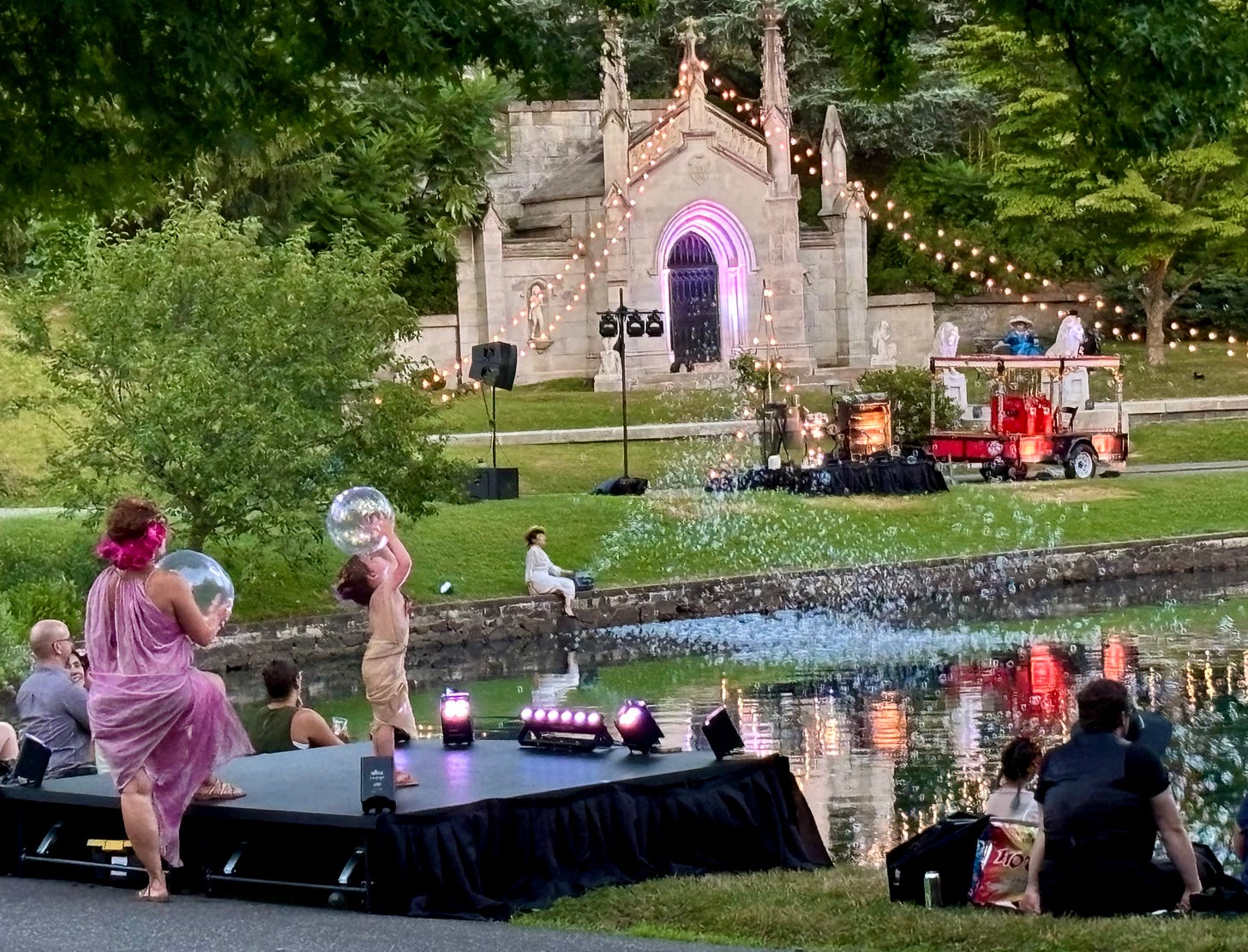 The Niblo's mausoleum, a grand stone structure draped in lights that hang over grass and connect to the edge of a lake. The arched entrance is bathed in purple light. On the shore of a light, a performer sits in an old-timey swimming costume running a bubble machine that sends flocks and flocks of bubbles floating over the water. On the shore closer the camera, a small stage is set up. A child in a ballet costume has a ball that looks like a bubble balanced on her head, with her hands on either side. Behind her, a woman in a lavender toga stands ready to join her, also holding a ball.