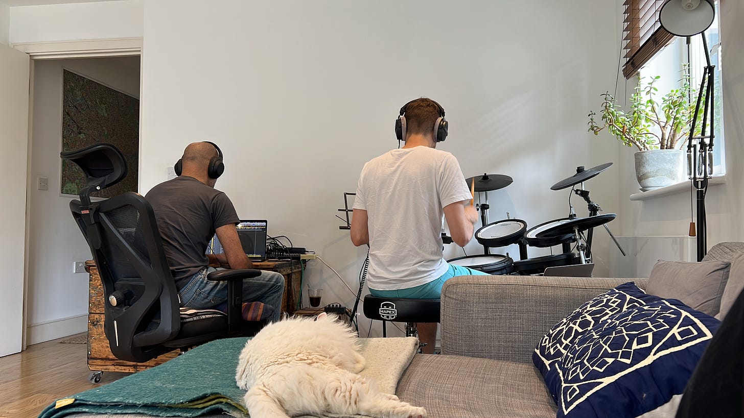 Two men sat down facing away from the camera. Imran is on the left on an office chair leant over a laptop. Jamie is on the right playing an electronic drum kit. A fluffy white cat is sleeping on the sofa in the foreground.