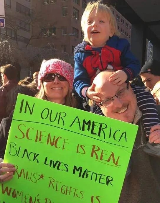 Image of a person holding a sign “In Our America: Science is Real, Black Lives Matter, Trans Rights is….” next to a man with a toddler on his shoulders. They are at the Women’s March. Photo courtesy of Jennifer Rogers-Brown.