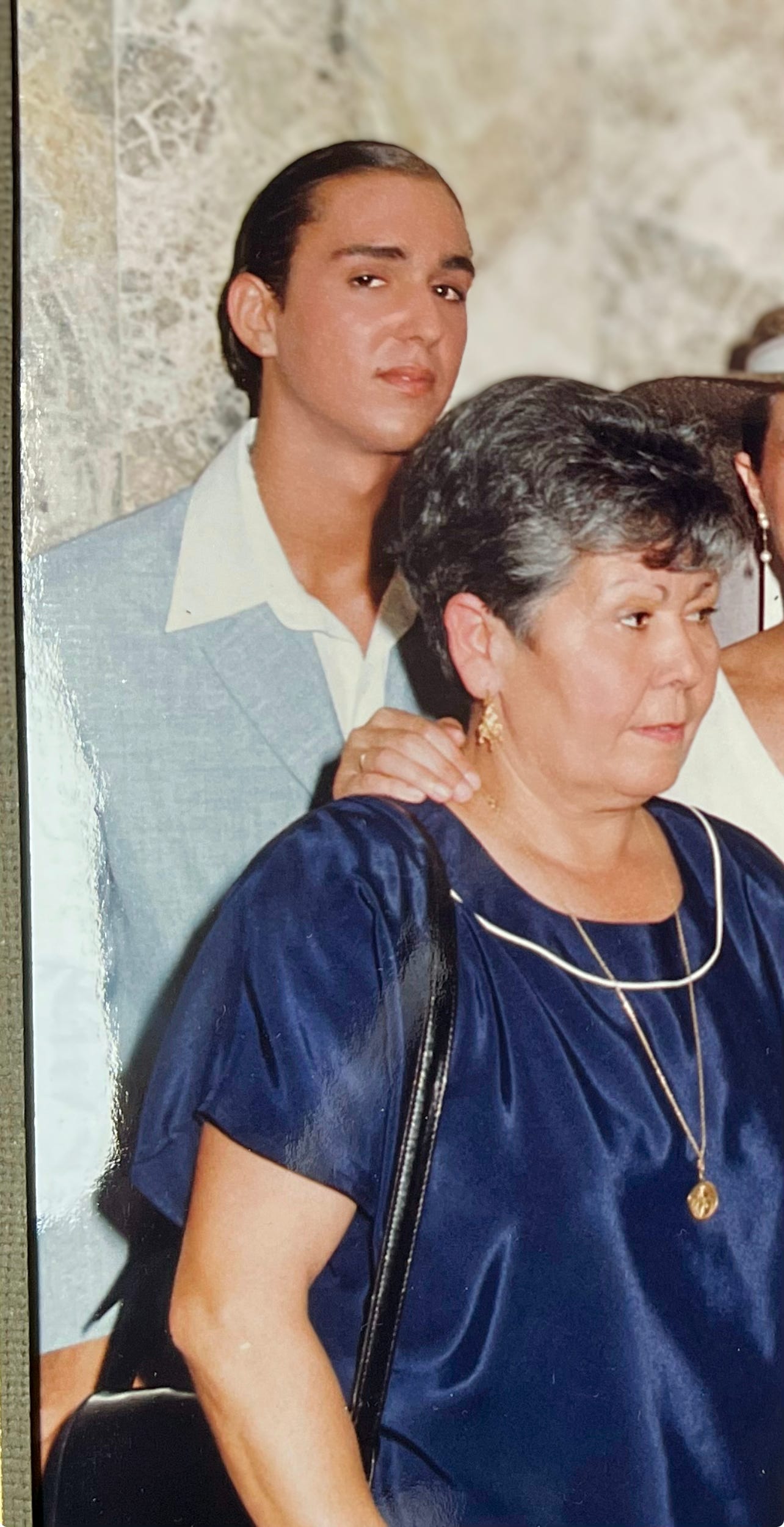 The image shows a vintage photo of myself and my mum captured in a close, affectionate stance at my sister’s wedding. I was 17, with slicked-back hair, a light blue shirt, and a prominent, confident gaze. My mum is dressed in a dark blue outfit, adorned with a gold necklace and matching earrings, exuding a look of composed matriarchal warmth. 