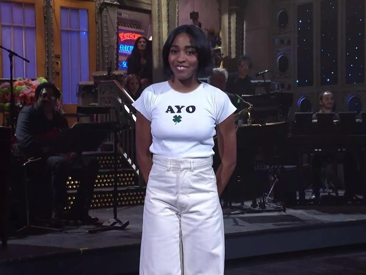 Ayo Edebiri, uma mulher negra retinta, na casa dos 20 anos, sorri para a câmera. Ela está no cenário do programa Saturday Night Live. Tem cabelos curtos e usa uma camisa que diz AYO, na fonte Cooper Black, típica dos anos 70. Abaixo do nome AYO, há um trevo de quatro folhas irlandês.