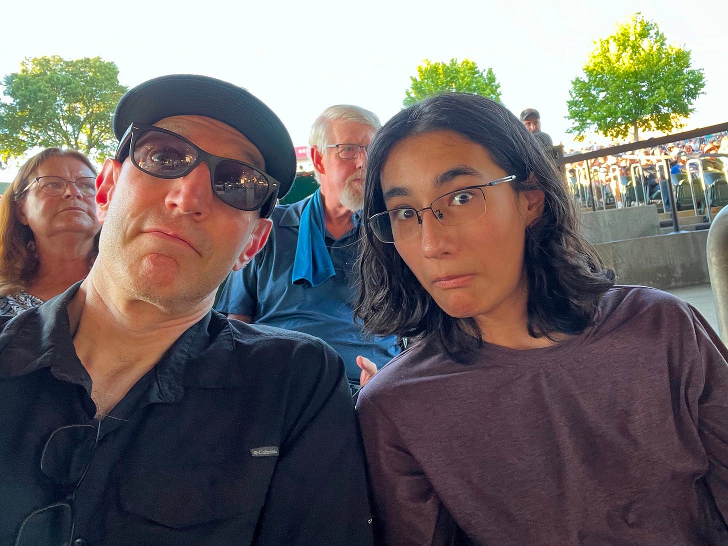 The author, left, a middle-aged man in a baseball hat, and his son, a teenager with long hair, mug for a selfie with other concert attendees behind.