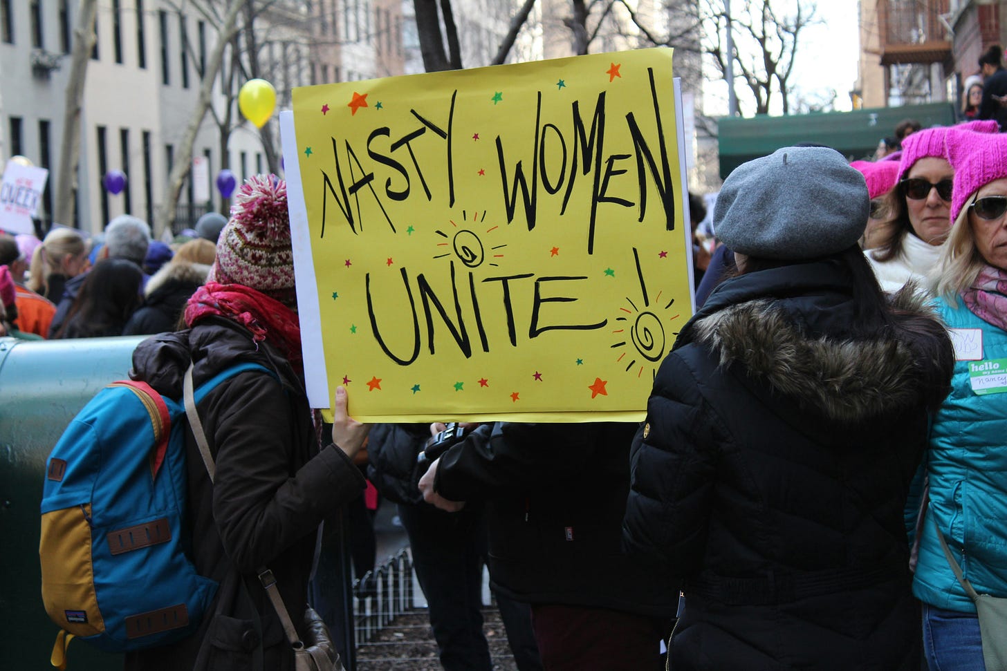 Taken from Women's Protests in New York. Women hold up a handmade sign that says "nasty women unite", as other women walk by wearing pink pussy hats. 