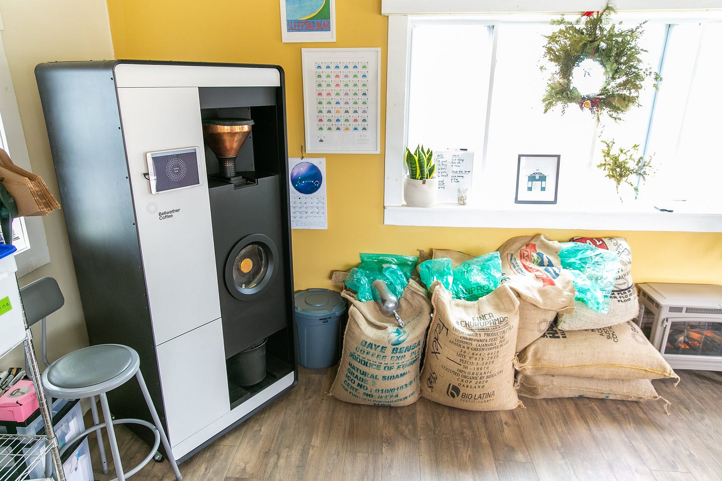 An electric coffee roaster that looks like a modern vending machine sits in the corner against a sunflower yellow wall. Burlap sacks of coffee beans are leaning up next to the machine.
