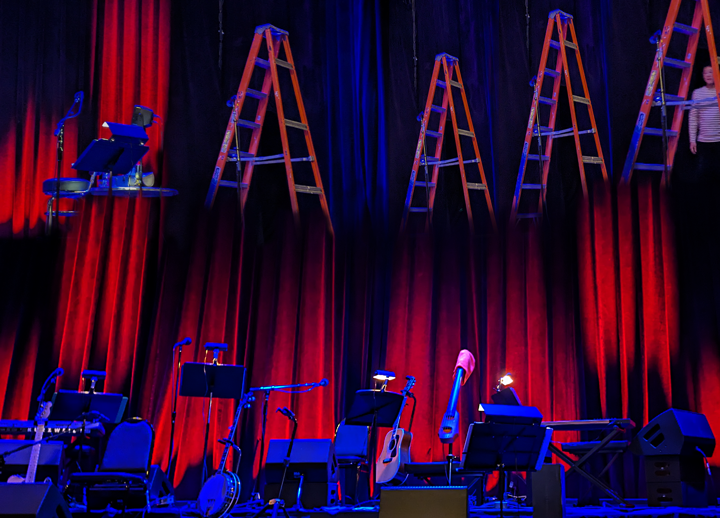 How I remember the Magnetic Fields at Town Hall in New York City. The stage is set with instruments waiting. Red curtains ascend, turning into ladders or disappearing into black or blue. A man in a striped shirt, blurred, peeks under one such ladder. A desk, unfinished, with microphone, floats.