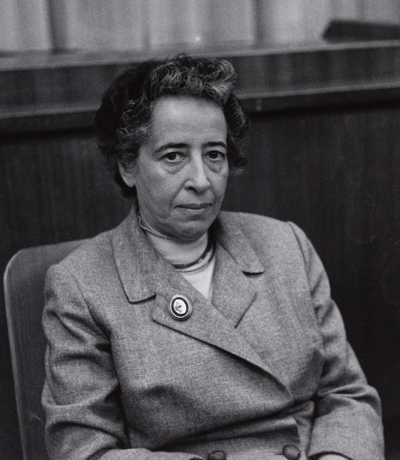 A stern black and white picture of Hannah Arendt, a slim, Jewish woman. She is wearing a masculine style tweedy looking suit with a brooch on the lapel. She has short, wiry, curly hair, fairly well cropped and greys surrounding her temple. She has striking deep, dark eyes that stare at the viewer almost defiantly.