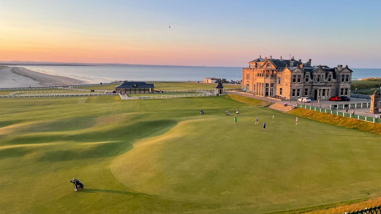 Aerial view of the St Andrews Old Course at sunset, showing the iconic clubhouse, expansive green fairways, and the West Sands beach in the background.