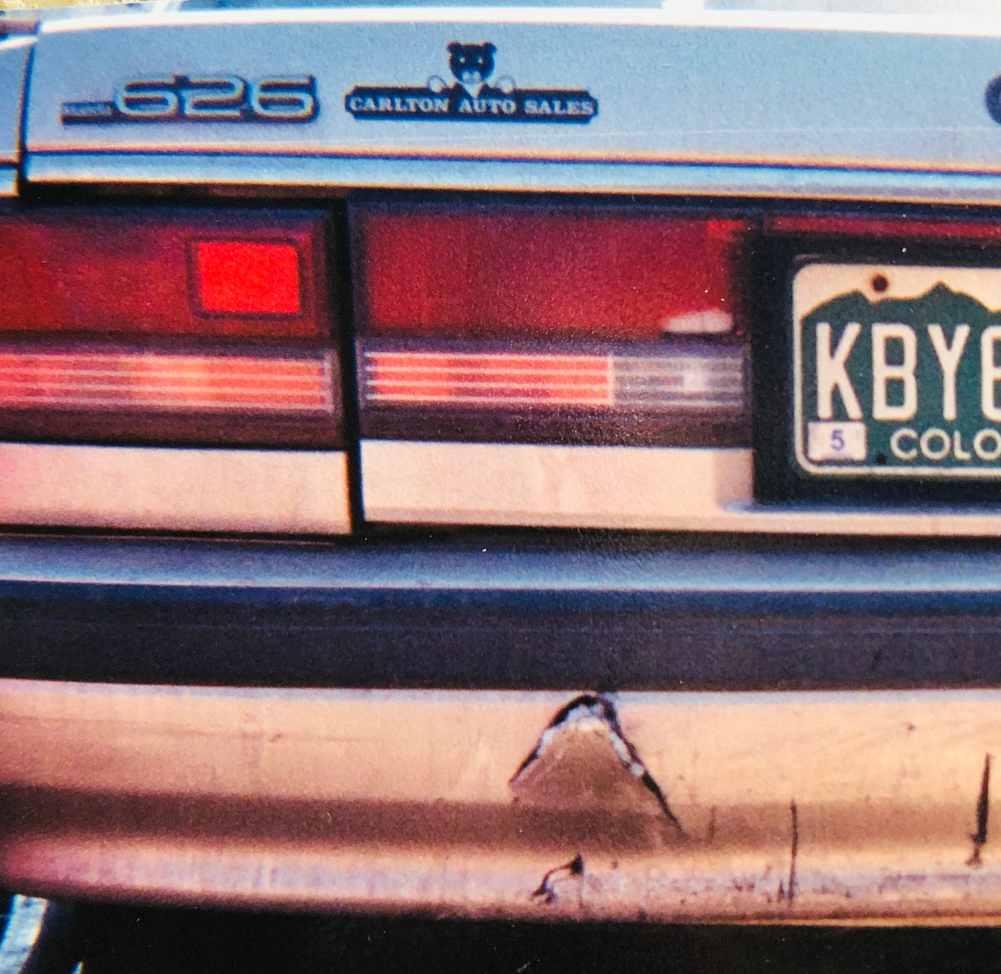 Back end of a Mazda 626 with Colorado plates--a triangle-shaped hole has been gouged from the bumper, along with a bunch of scrapes.