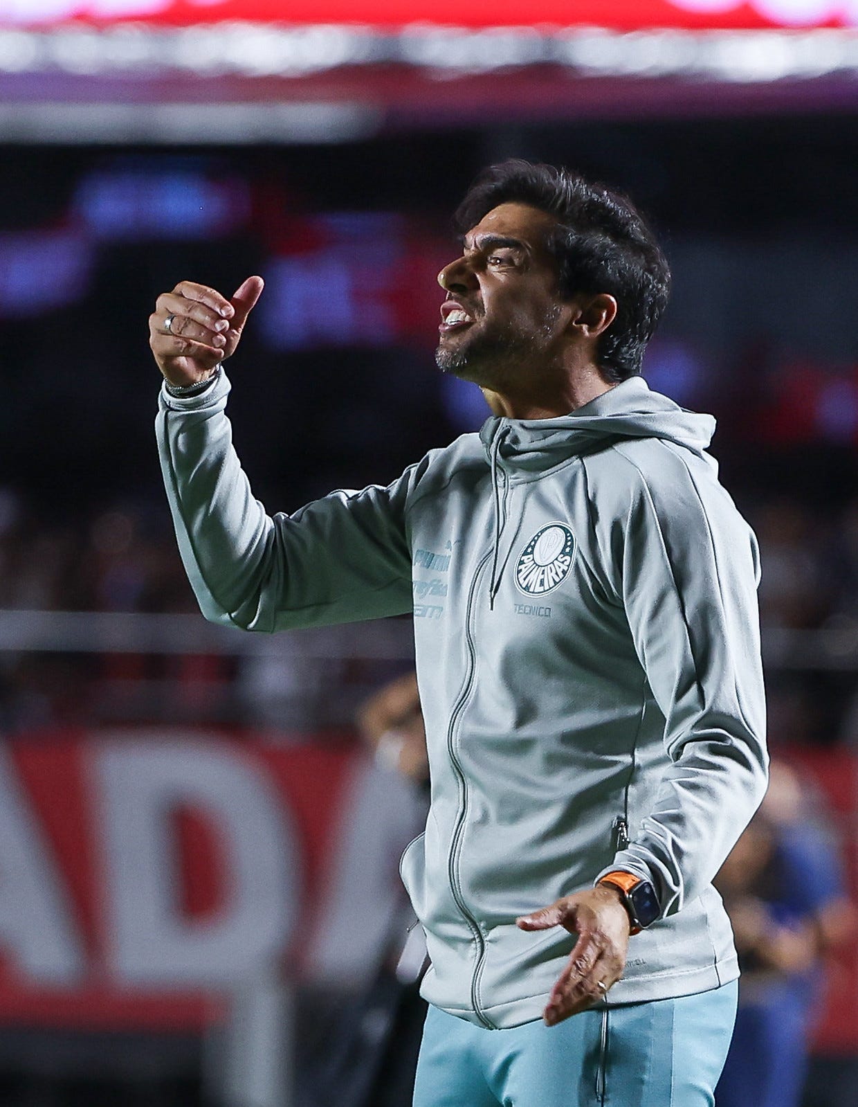 Abel Ferreira durante o jogo São Paulo x Palmeiras. Ele está de perfil, olhando da direita para a esquerda, com calça verde clara e casaco cinza, com os dentes rangidos e a mão direita levantada em sinal de reclamação