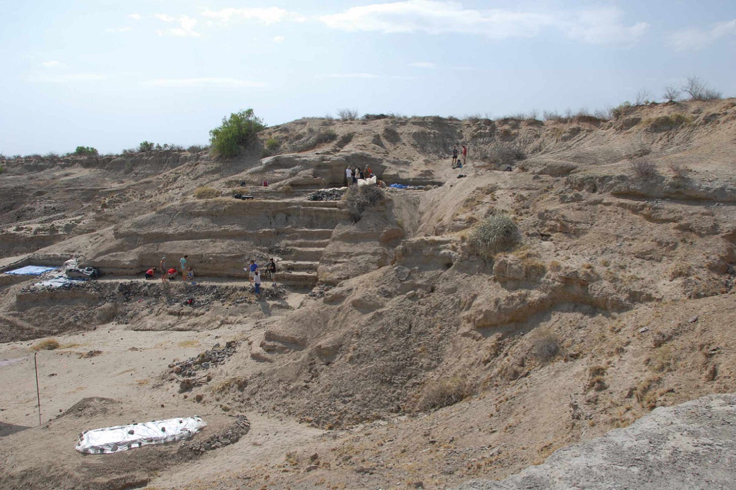 Tan hills with little vegetation, one heavily altered by digging a series of flat surfaces along its slope. Archaeologists stand on some of these surfaces. Tan hills with little vegetation, one heavily altered by digging a series of flat surfaces along its slope. Archaeologists stand on some of these surfaces.