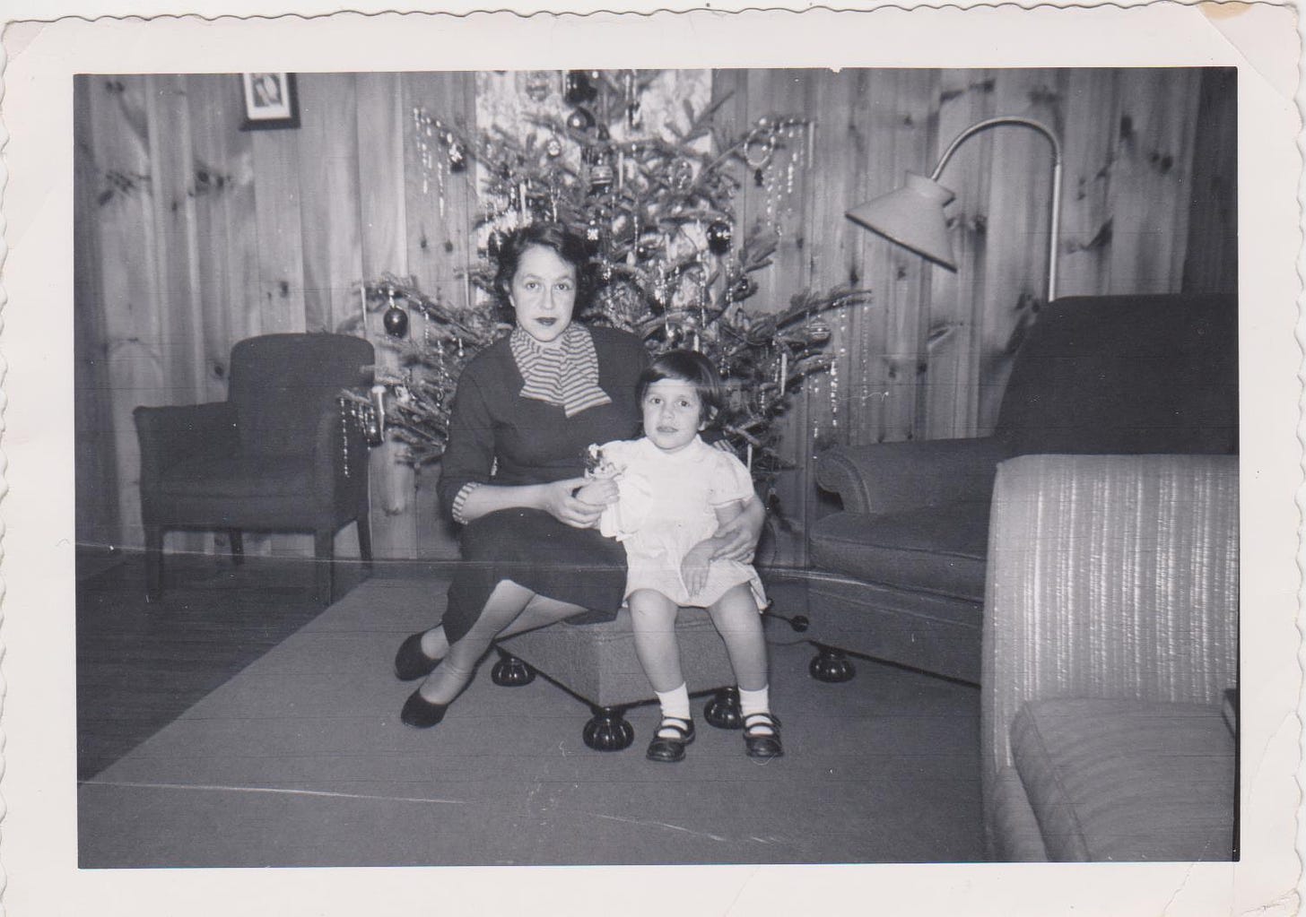 A little girl in a white dress and Mary Janes sits next to her mother in front of a Christmas tree.