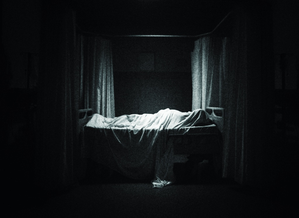 A figure sleeps underneath a sheet in a hospital bed in a dark room