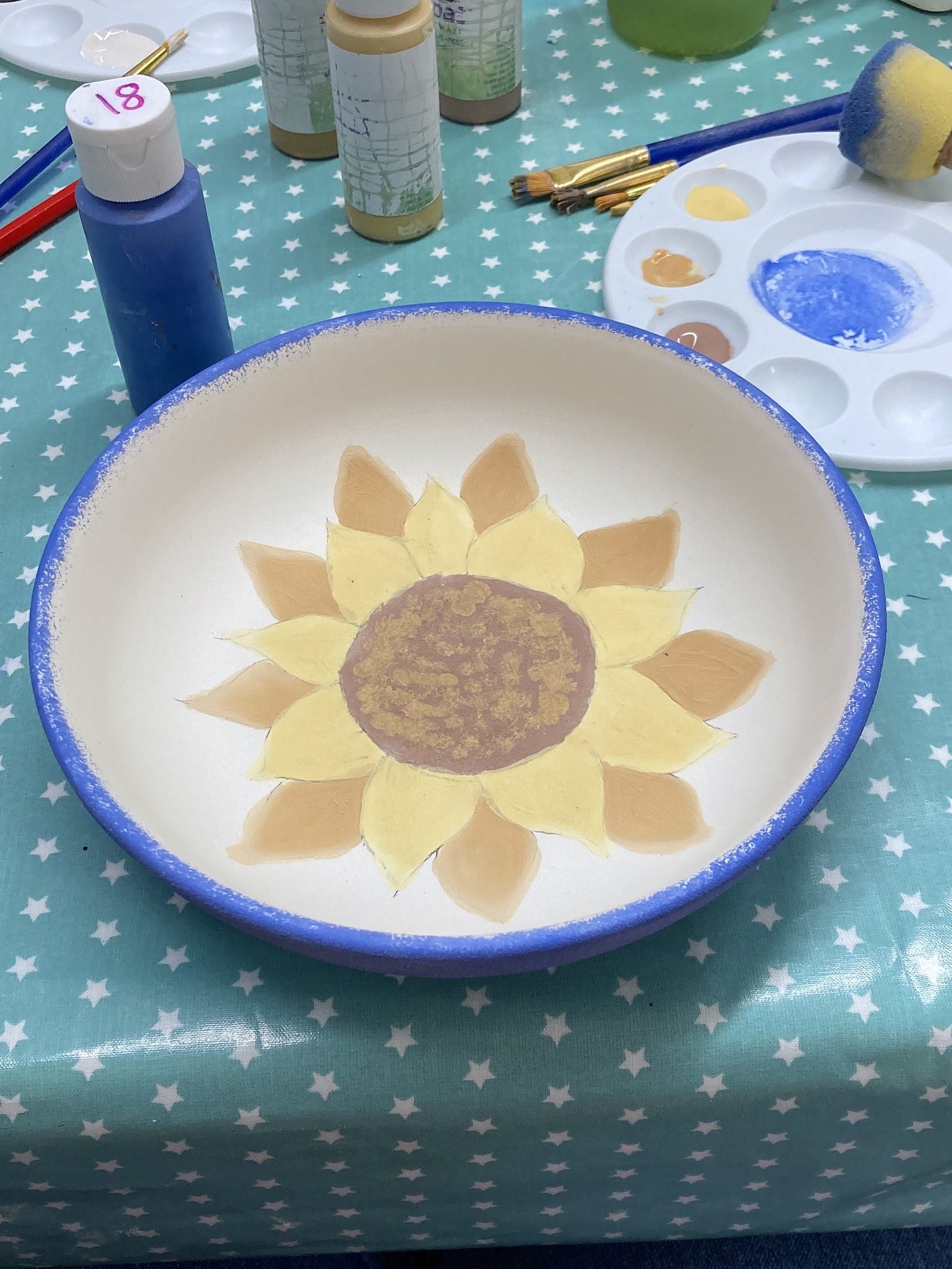 Pottery painting table. In the centre of the image is a white bowl with a sunflower painted inside and a sponged royal blue rim and outside.
