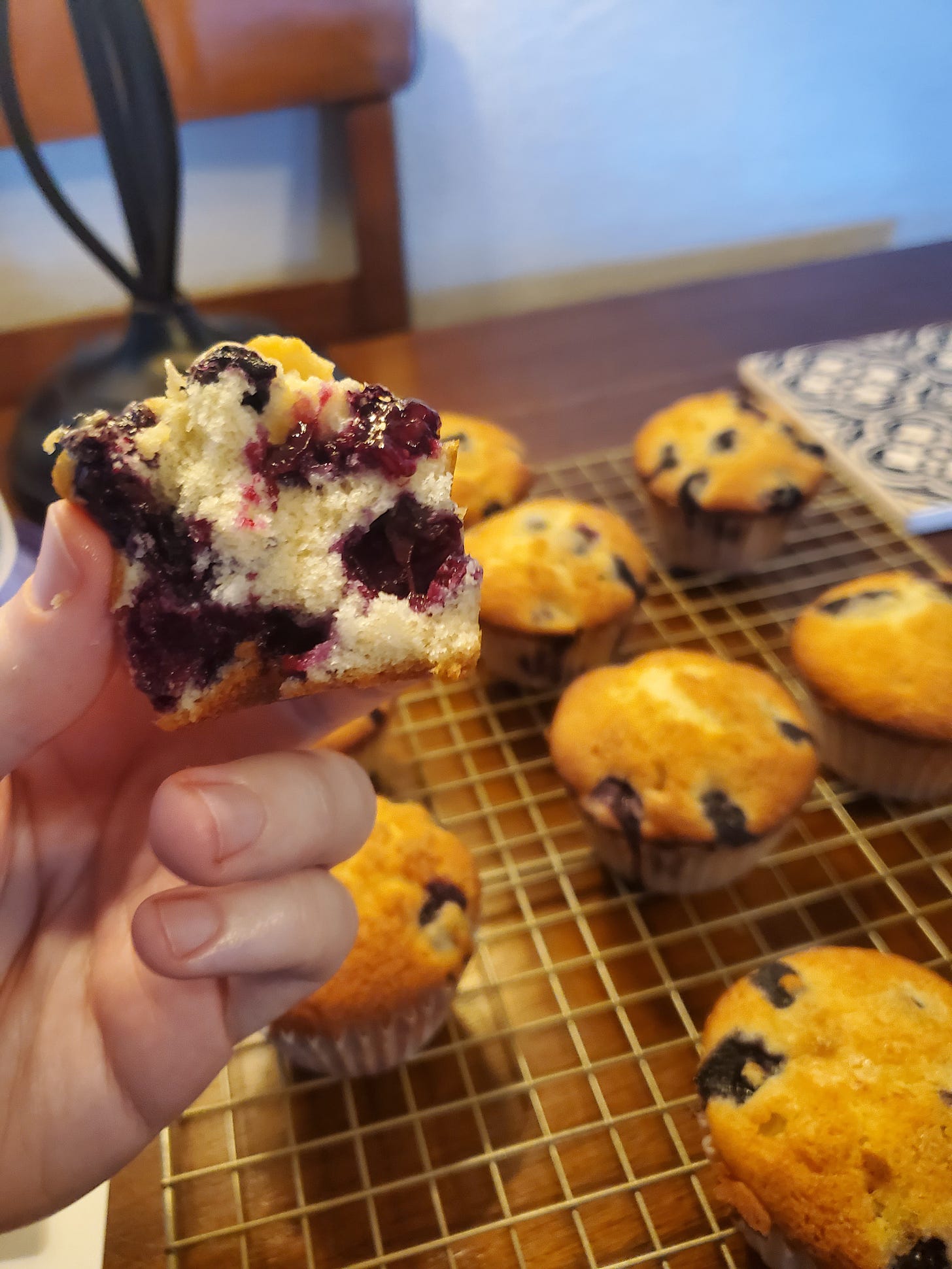 A close up image of half of a blueberry muffin. Other muffins sit on a golden cooling rack in the background, out of focus.