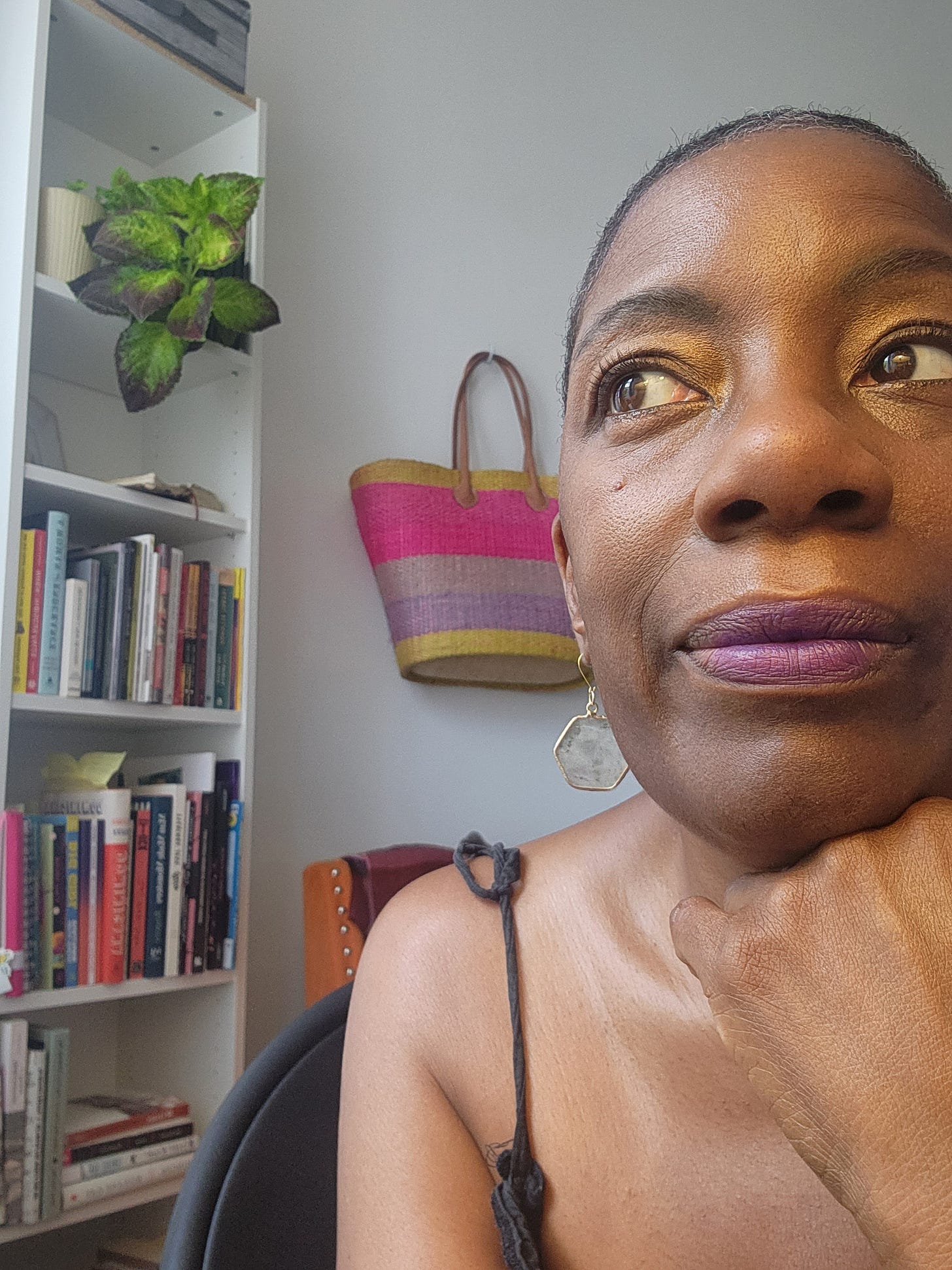 Black woman with shaved close hair in sun filled room. To the left is a white book shelf, with a very green plant at the top and a slim shelf. A basket with a beige handle in block colors of beige, pink, and purple hangs on the wall just behind Anna.
