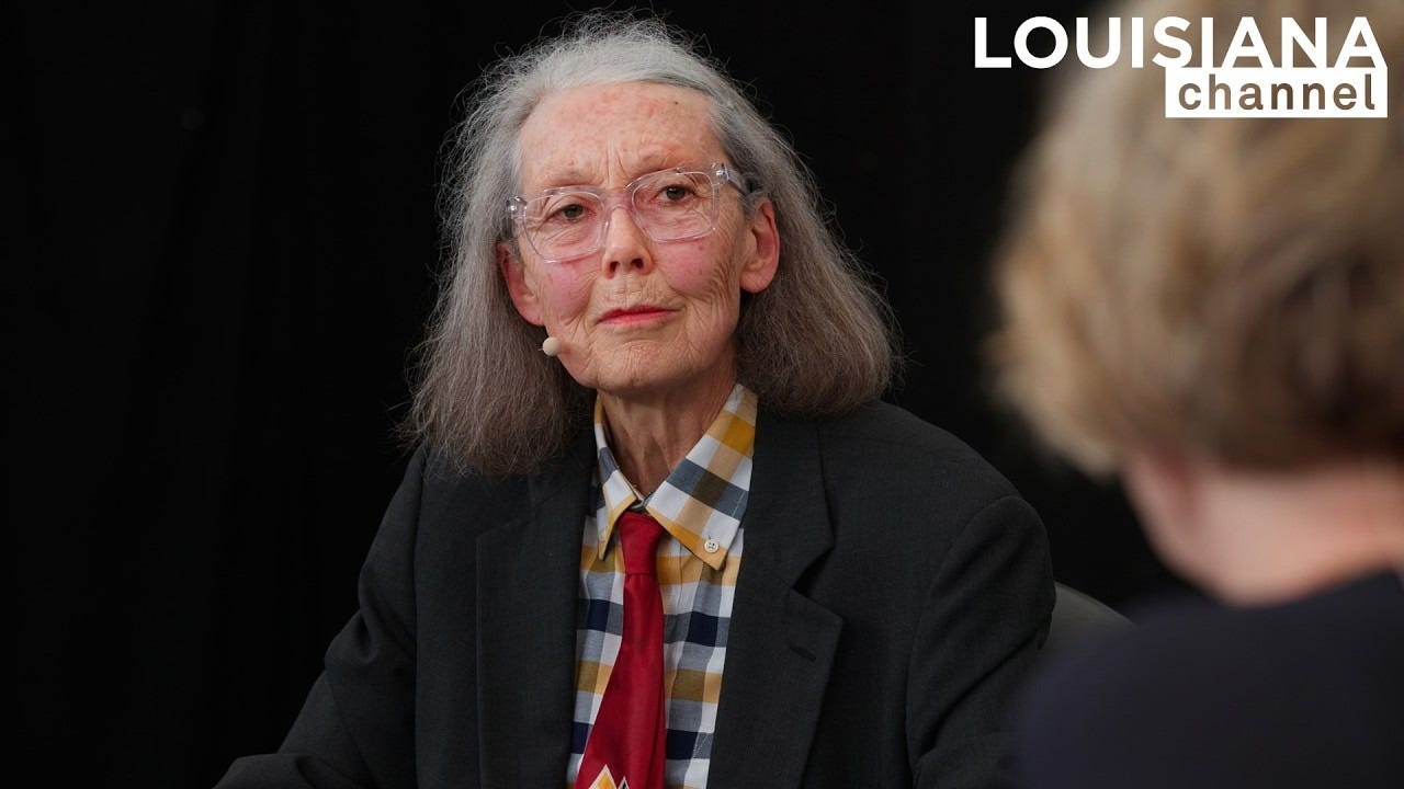 A screenshot of Anne Carson who has long gray hair, glasses, a dark suit, and a red tie, sits in a discussion setting with a black background with the Louisiana Channel logo in the corner. A screenshot of Anne Carson who has long gray hair, glasses, a dark suit, and a red tie, sits in a discussion setting with a black background with the Louisiana Channel logo in the corner.