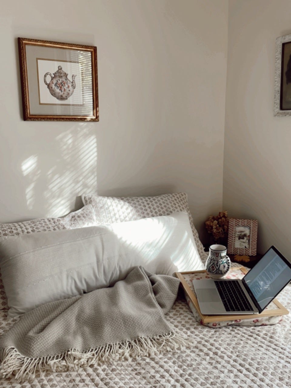 A bed with a pink quilt has an open laptop, blanket, and mug resting on top. A picture of a teapot is hanging on the wall.