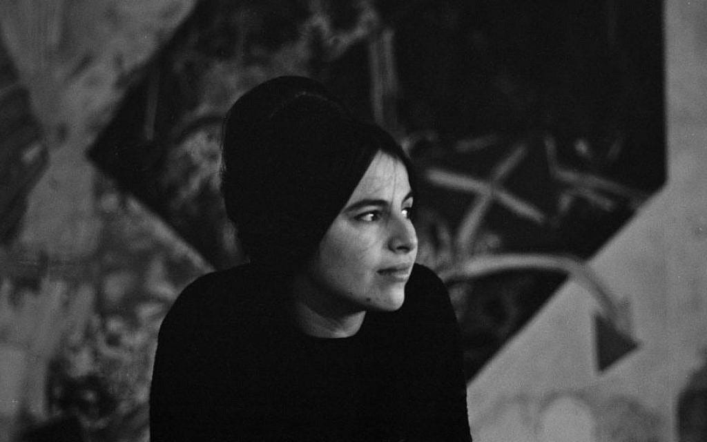 Black and white photograph of a young Eva Hesse with her black hair in an updo, gazing to one side. There is a blurred work of art behind her.