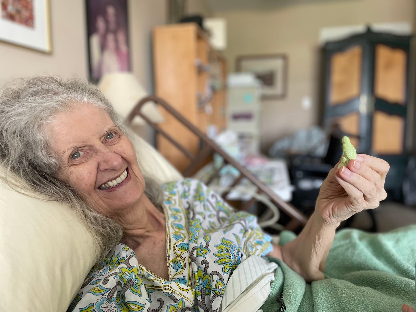 An elderly woman -- my mom -- smiling from a hospital bed set up in an apartment. She holds a small, green porcelain bird.