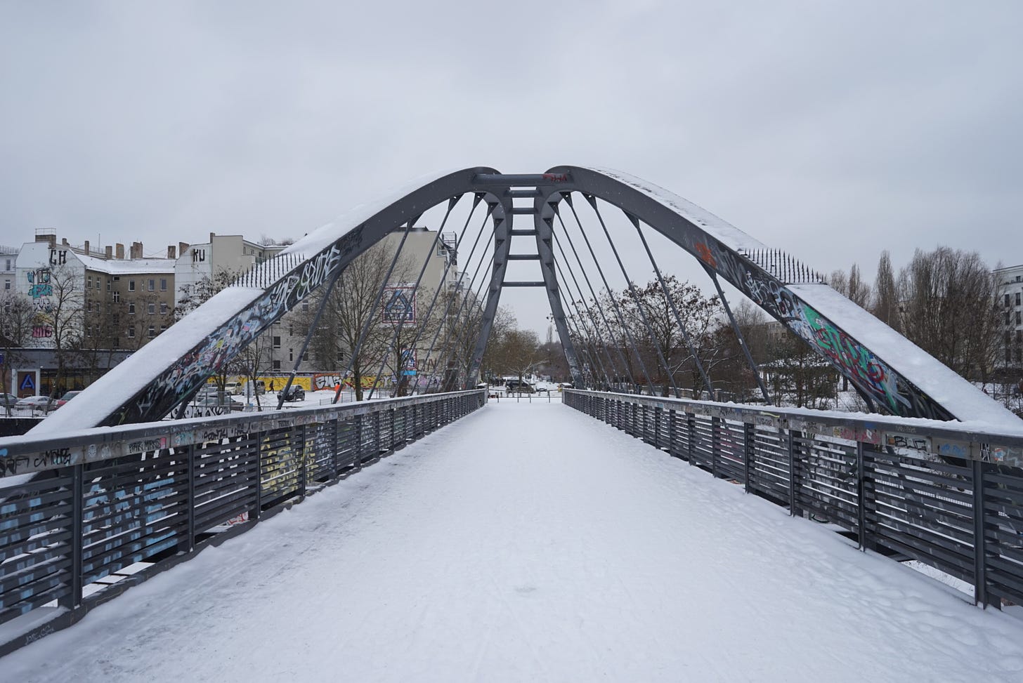  Imagem de uma ponte arqueada coberta de neve com corrimões metálicos grafitados. Edifícios ao fundo sob um céu nublado.