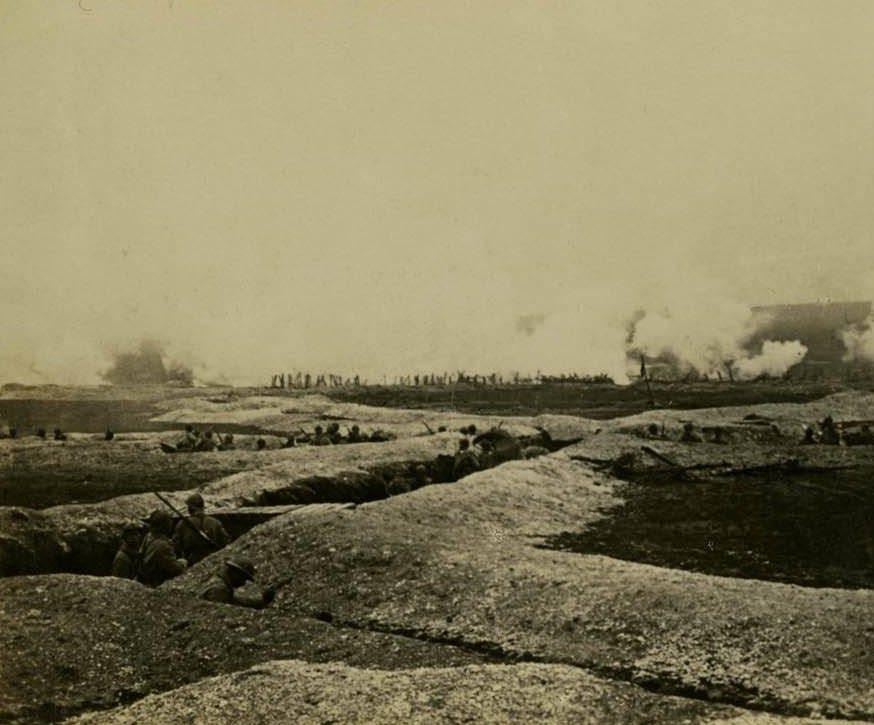 A photograph, taken from one half of a stereograph image card, of a German Attack in North Compiegne, France. Troops can be seen in trenches in the foreground, smoke envelopes the battlefield further away.