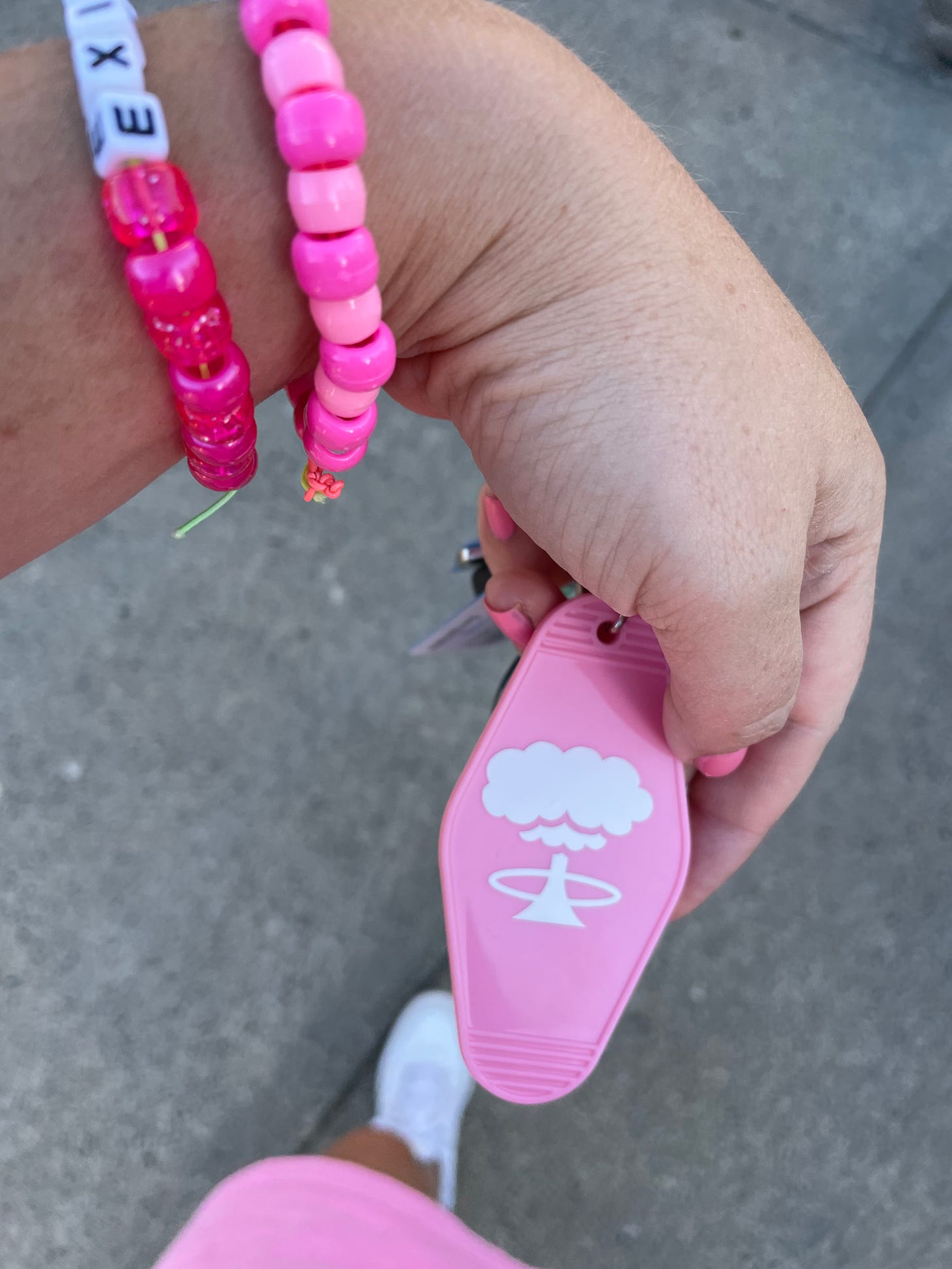 a white hand with pink beaded bracelets on it is holding a pink plastic motel like keychain with a white mushroom cloud on it