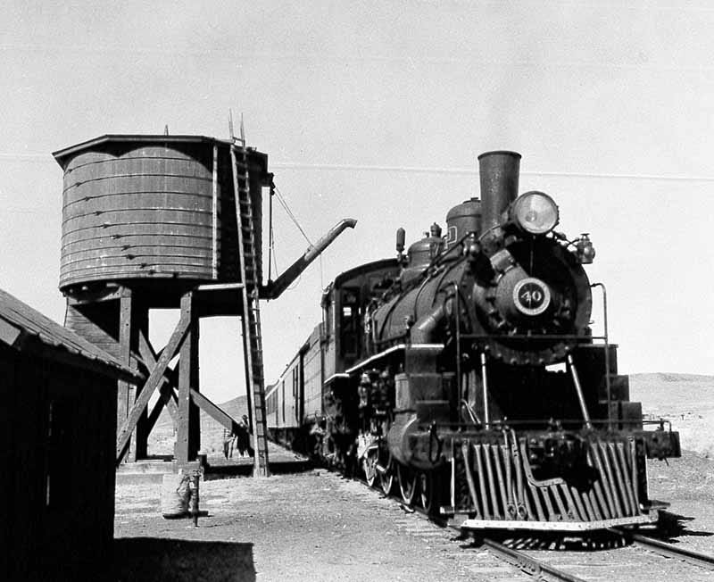 A black and white photo of a steam locomotive with cow catcher stopped on the railroad tracks besides a short, stout, wooden water tower. The water tower is built like a barrel with wooden slats held together by metal straps around the circumference. The water tower sits on short stout struts. A ladder runs from the base of the tower to the top. An articulated arm reaches from the tower to the steam train to deliver water.