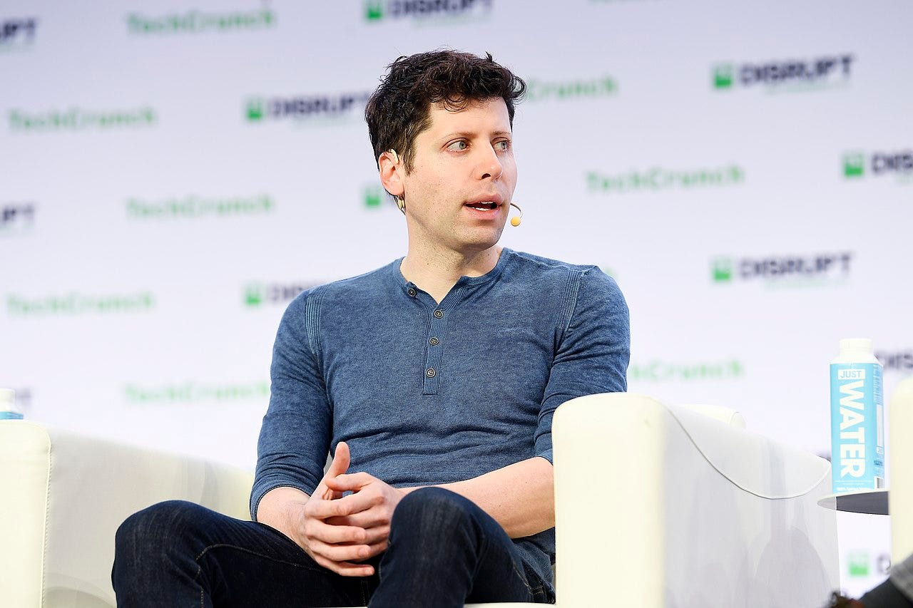 Sam Altman sits in armchair wear a microphone in front of a backdrop with blurry words "TechCrunch" and "Disrupt."