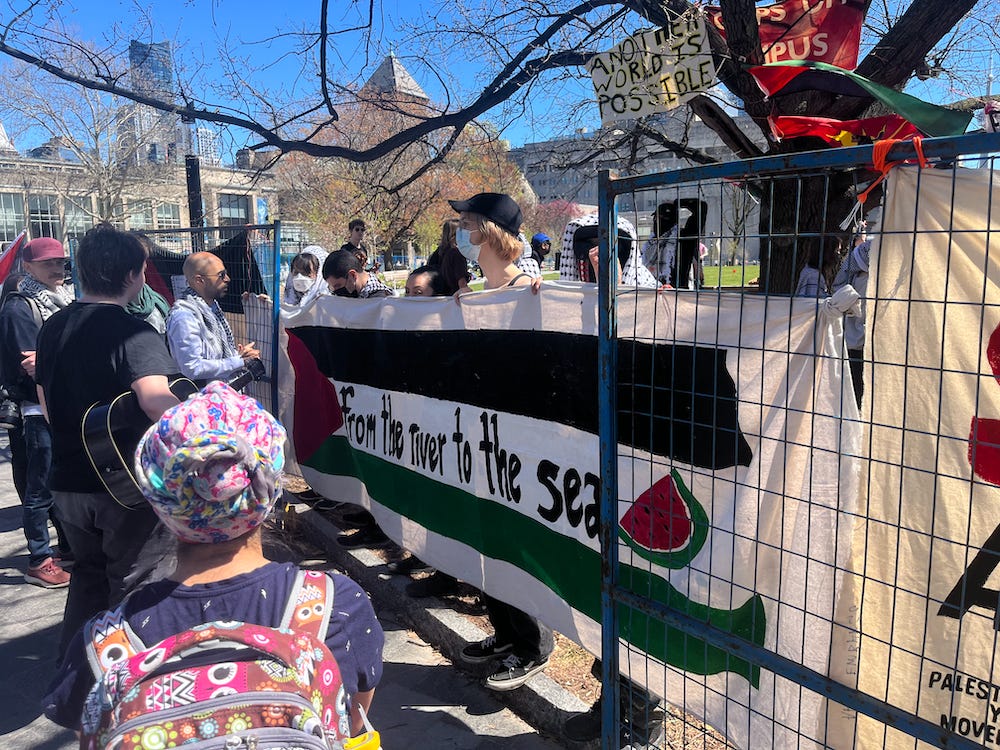 masked people hold a banner reading "from the river to the sea" in a fence opening