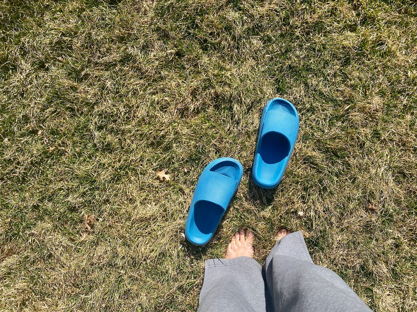 A landscape-oriented photograph of grass. In the bottom right, a person's legs in gray sweatpants and bare feet are viewable. In front of the feet are two medium blue slides sitting in the grass. The photo is of me while I'm grounding, also called earthing, but walking barefoot in grass