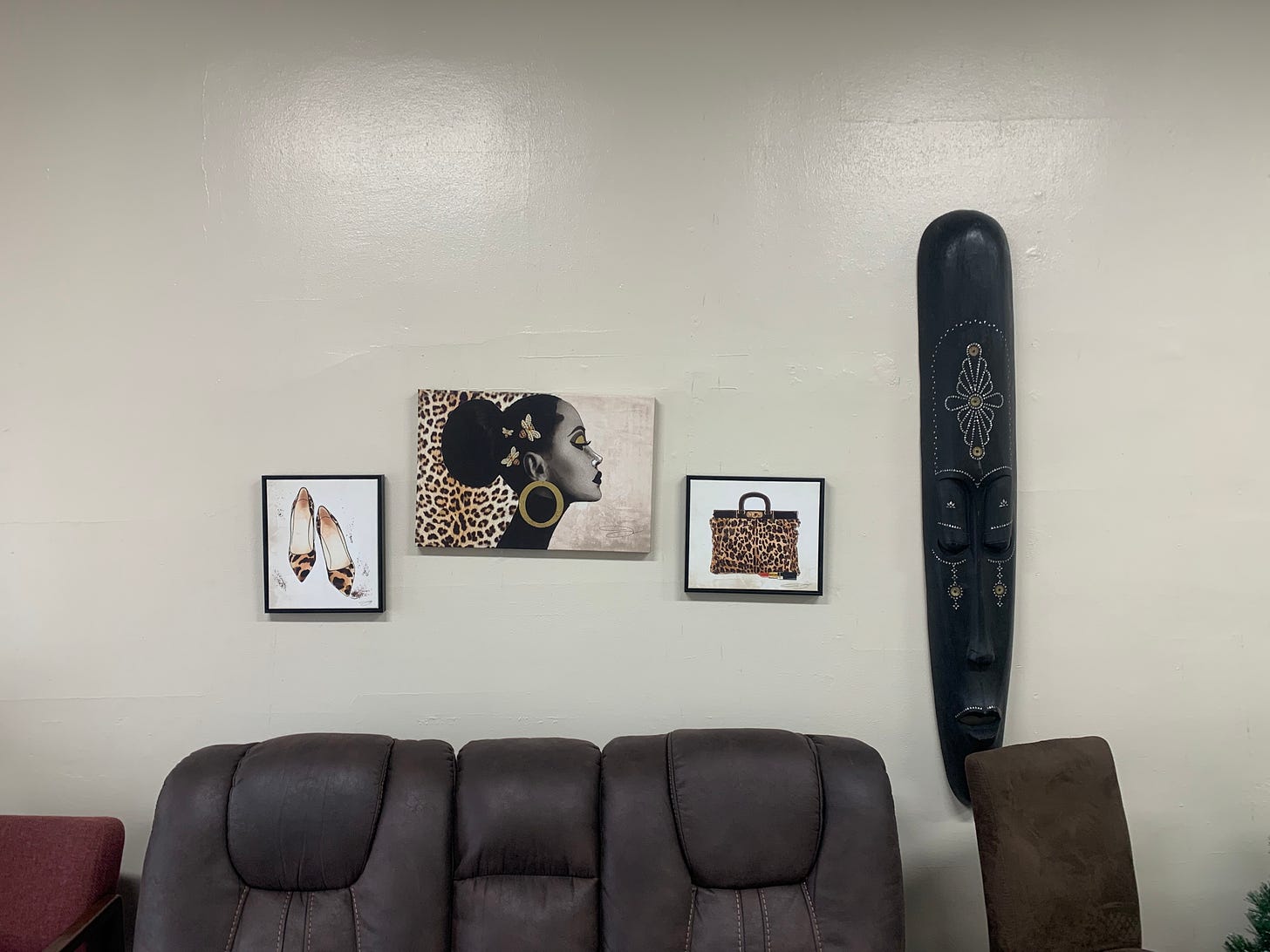 Three pictures, one of a  pair of slippers, one of a Black woman with hoop earrings, and one of a bag, hang above a couch. A black, elongated mask hangs beside the pictures.