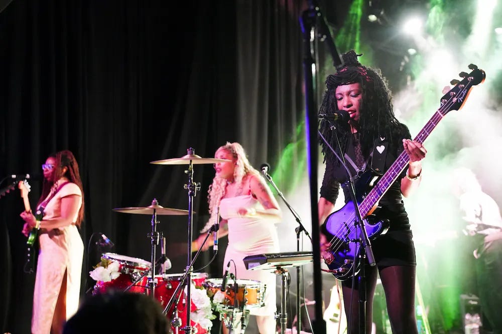 On a stage, three Black women stand in a line playing their instruments while standing at microphones. Far left is the guitarist in a white dress. In the middle, the drummer stands at her kit in a white dress. Closest to the camera is the bassist wearing all black and signing into her microphone.