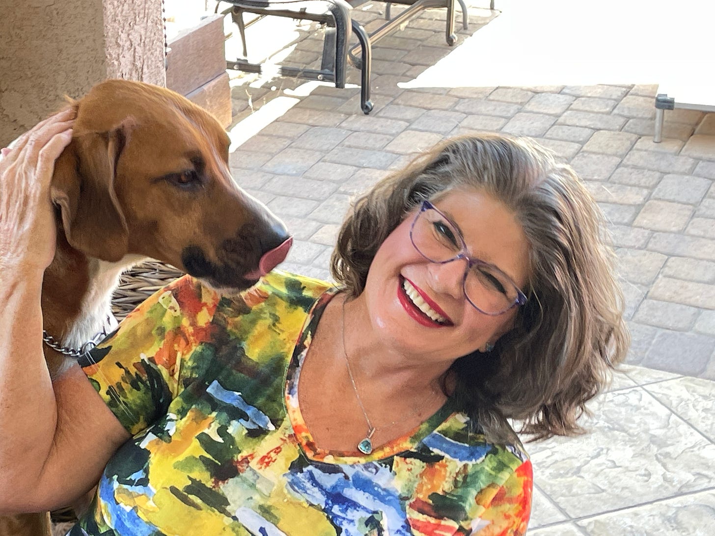  A large russet-colored dog with his tongue out next to a woman with a brightly-colored shirt , eyeglasses, and salt and pepper hair.