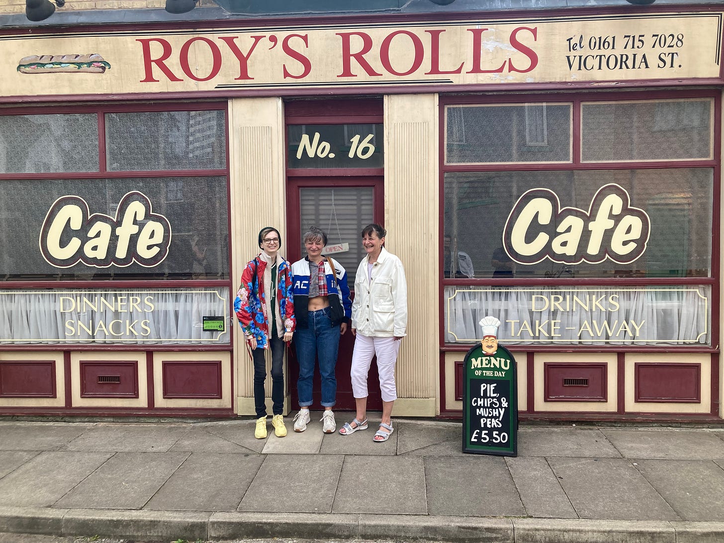 Photograph of Beck, Beck's auntie and Beck's mum standing on a pavement in front of Roy's Rolls cafe on the Coronation Street set. The windows advertise they sell dinners, snacks, drinks and take-away and there is a sandwich board advertising pie, chips and mushy peas are the menu of the day for just £5.50.