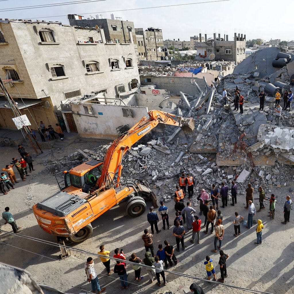 A vehicle works on damaged buildings as people stand on and near rubble.