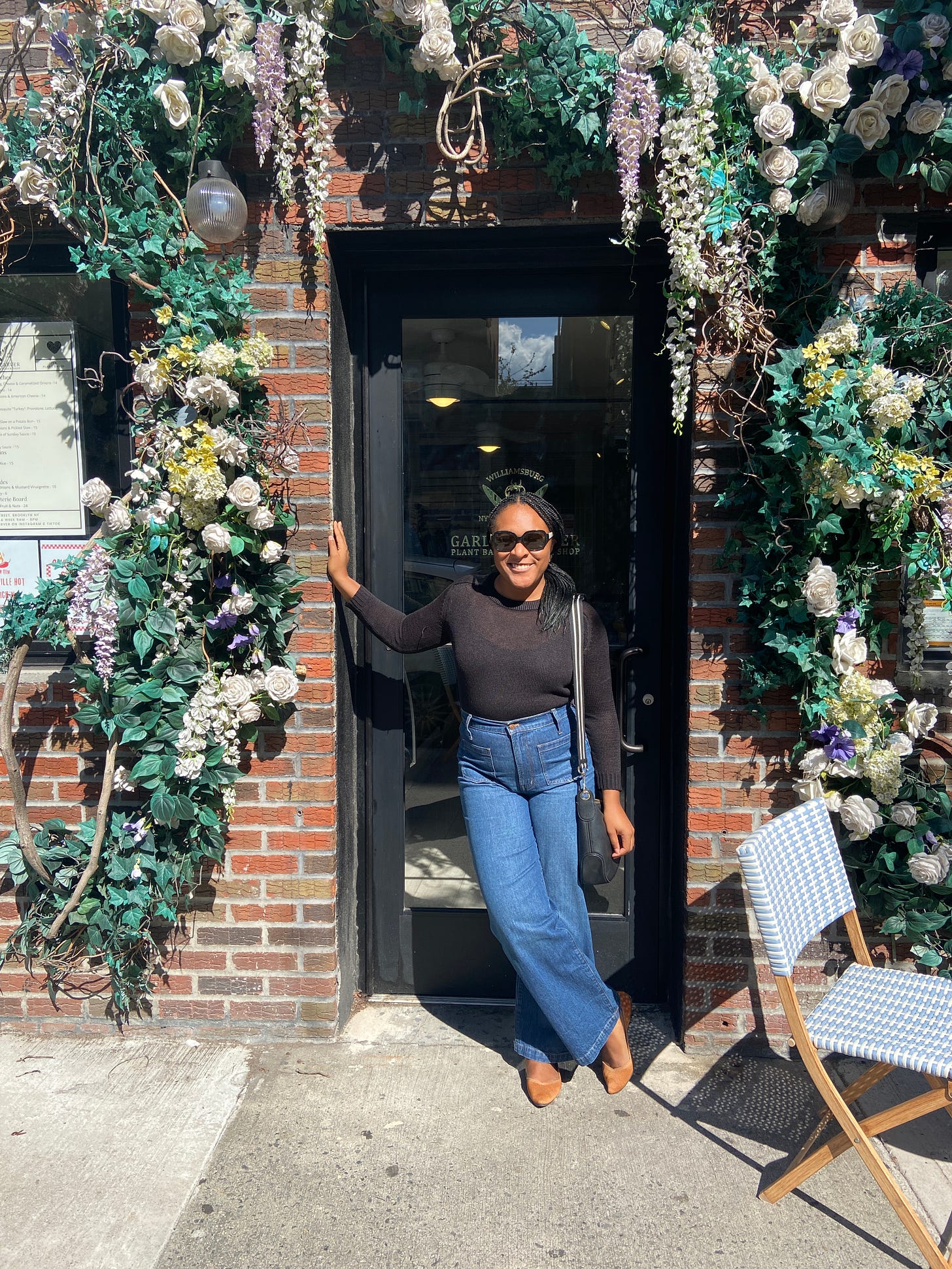 Alexa is leaning against a doorframe covered in a beautiful flower/leaf arch. The building is made of brick. Alexa is wearing a black sweater, flare blue jeans, and tan pointed flats. She has a black bag with a black and white striped strapped on her shoulder. She is wearing black sunglasses, and has her hair in a side ponytail. She is smiling at the camera.