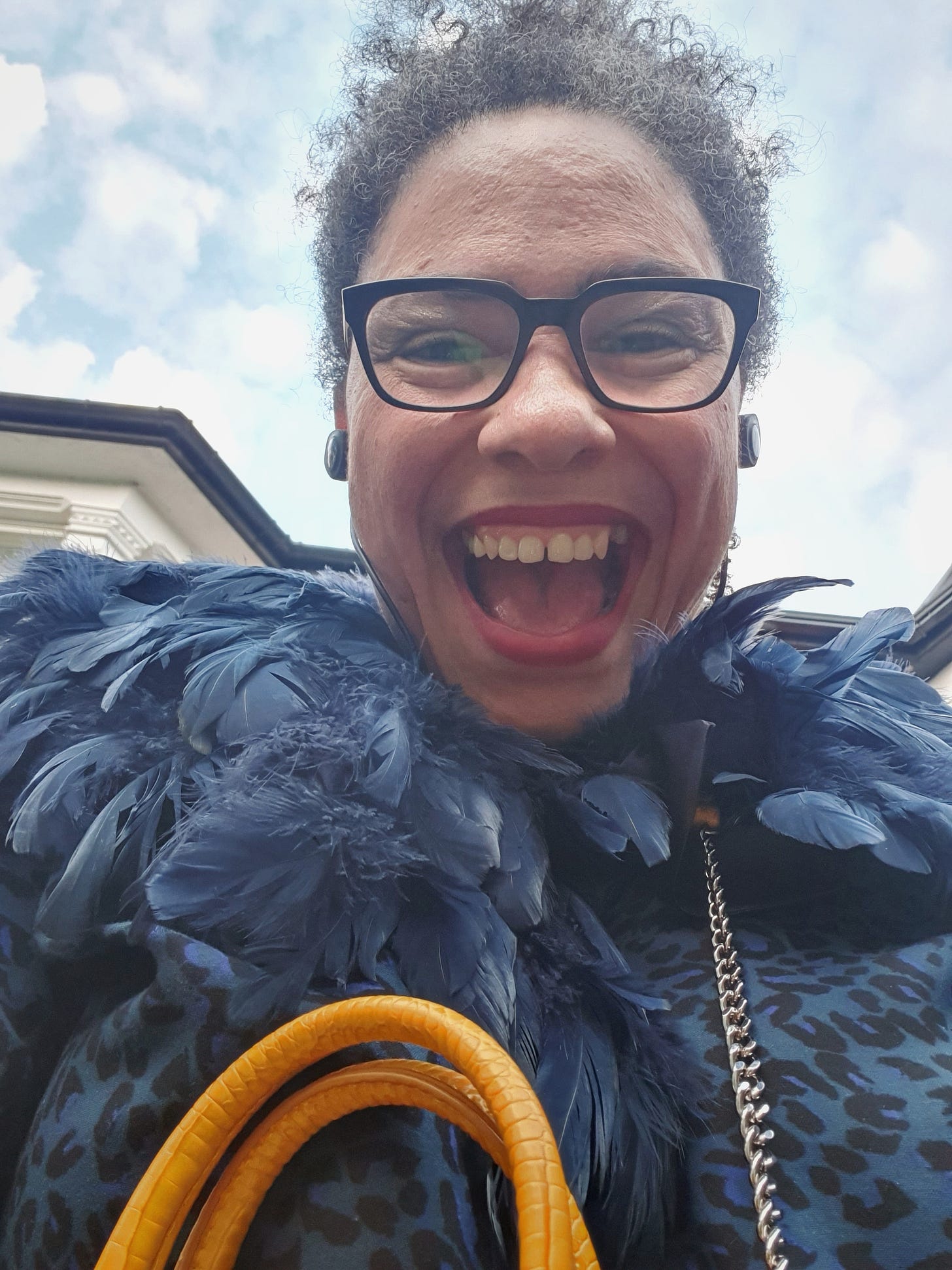 Photo of a woman looking down into the camera with the sky above, she is wearing glasses and a blue feather collar with a blue leopard print top