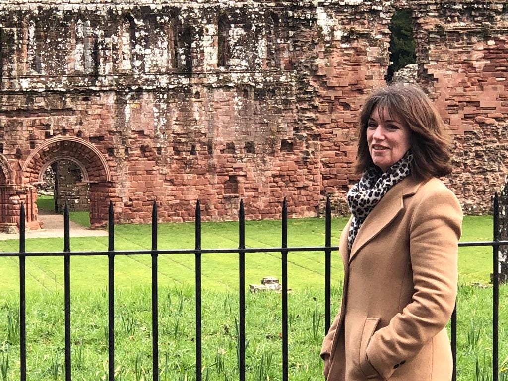 A woman with shoulder length brown hair and wearing a camel coloured coat stands smiling in front of an iron railing fence. Behind her is a large ruined ancient building made of sandstone, with a fancy archway to the left of the picture.