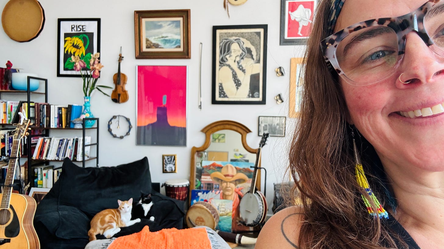 lisette in the foreground with lots of art hanging on the walls with instruments scattered around and two cats on a chair in back.