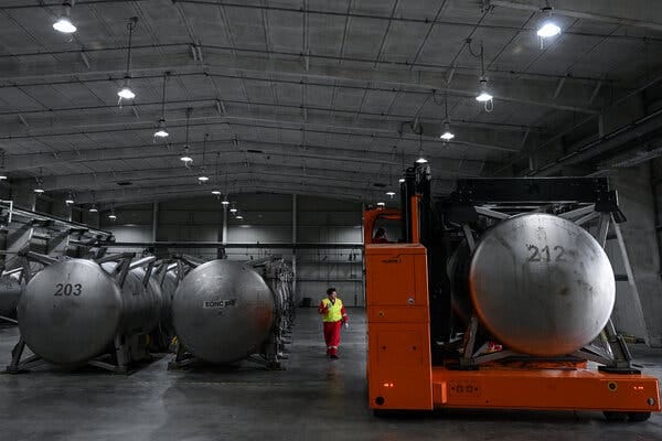 A man wearing a red jumpsuit and yellow vest walks in a large warehouse filled with rows of giant metal cylinders that are taller than the man. One cylinder is loaded onto an orange forklift.